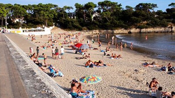 Ein Strand mit Menschen, die sonnenbaden und schwimmen, ein Boot auf dem Wasser und eine felsige Küstenlinie.