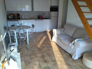 A compact kitchen with white cabinets, a dining table with chairs, a sofa, and a wooden ladder. - House in private hamlet near the beach (Vaux-sur-Mer)