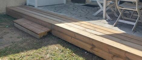 Een terras met een houten terras, een witte bank, een tafel met stoelen en een schuifdeur van glas.