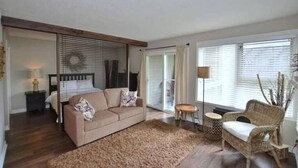 A living room with a beige sofa, a small round table, a wicker chair, and a large window with blinds. - Ski In Ski Out Cozy Studio (Blue Mountain)