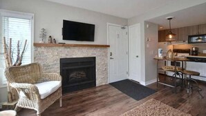 A cozy living room with a fireplace, a wicker chair, a television mounted on the wall, and a kitchen area with bar stools. - Ski In Ski Out Cozy Studio (Blue Mountain)
