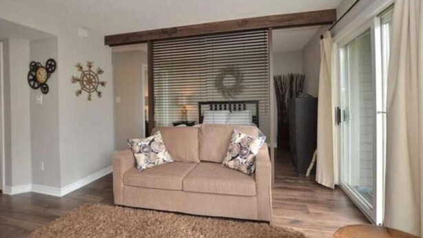 A living room with a beige sofa, a wooden ceiling beam, and a large window with blinds. - Ski In Ski Out Cozy Studio (Blue Mountain)