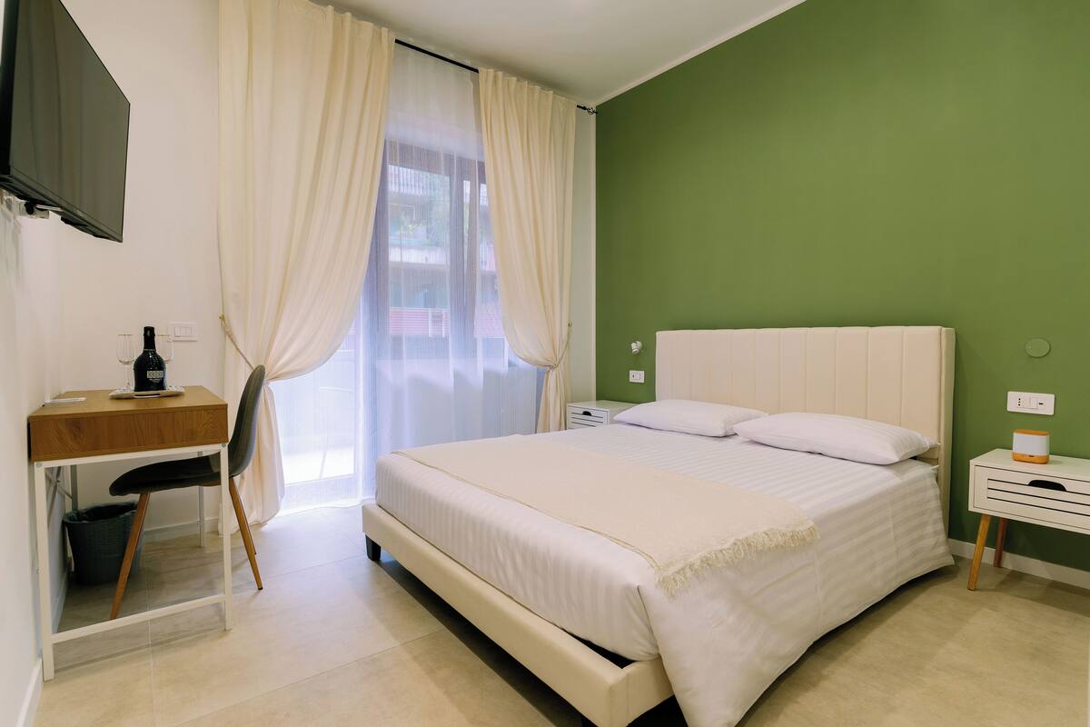 A bedroom with a bed, a desk, a chair, a TV, and a window with curtains.