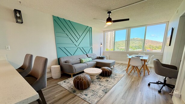A modern living room with a sofa, coffee table, and chairs. There is a dining area with a table and chairs. The room has large windows with a view of buildings and hills.