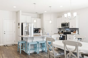 A modern kitchen with white cabinets, a central island with stools, a dining table with chairs, and pendant lighting.