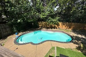 A backyard with a swimming pool surrounded by a brick border, a wooden deck, and a grassy area.