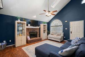 A living room with a fireplace, a ceiling fan, and a large window.