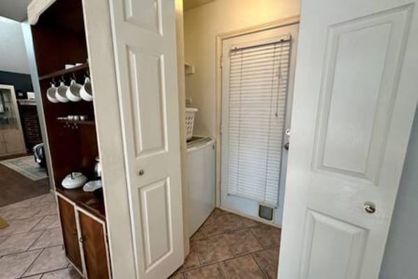 A hallway with a white door, a window with blinds, and a kitchen area with cabinets and dishes.