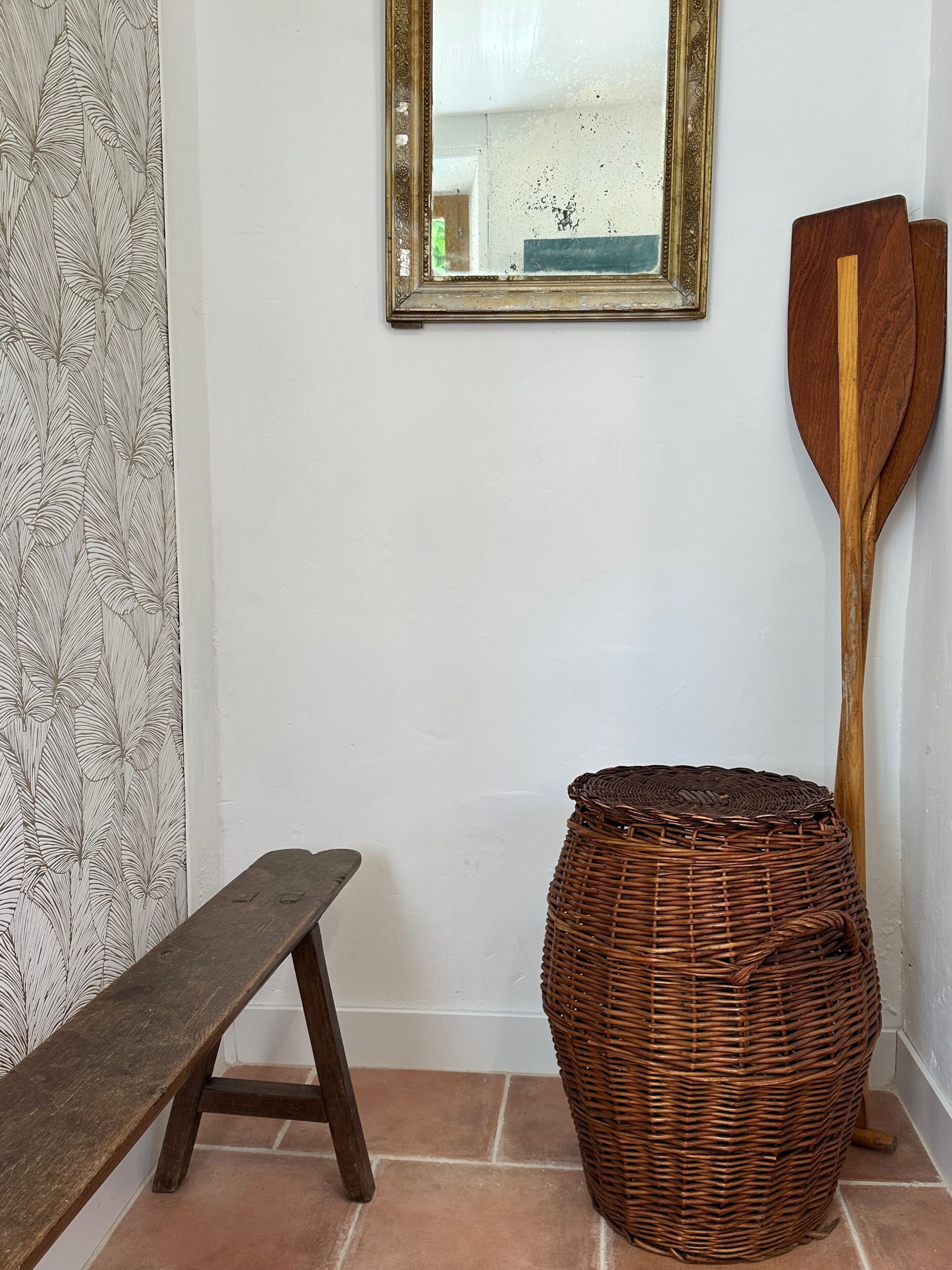 Une pièce comprenant un banc, un panier en osier et une pagaie en bois appuyée contre un mur orné d’un miroir.