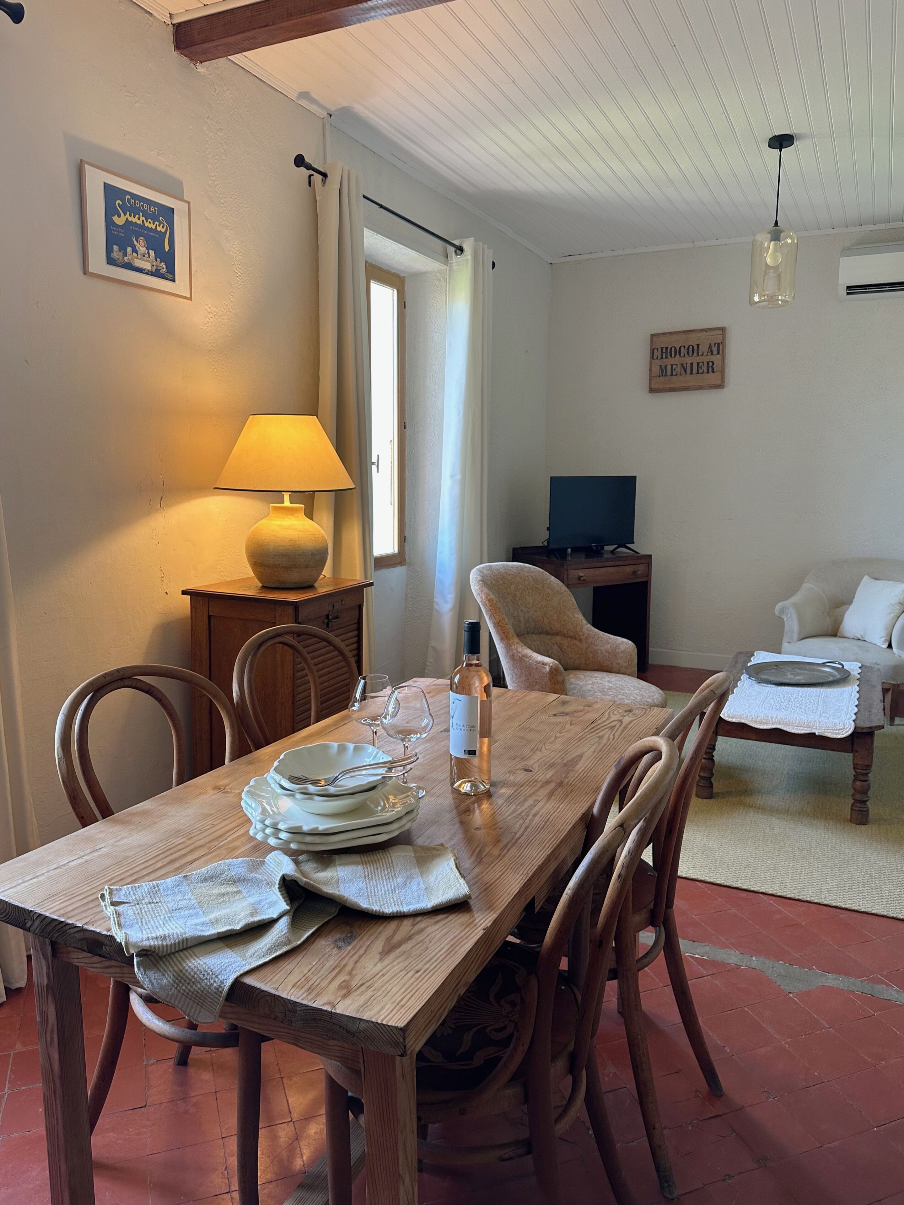 Un coin repas avec une table en bois dressée pour quatre personnes, une lampe et une bouteille de vin. On aperçoit un téléviseur et un canapé au fond.