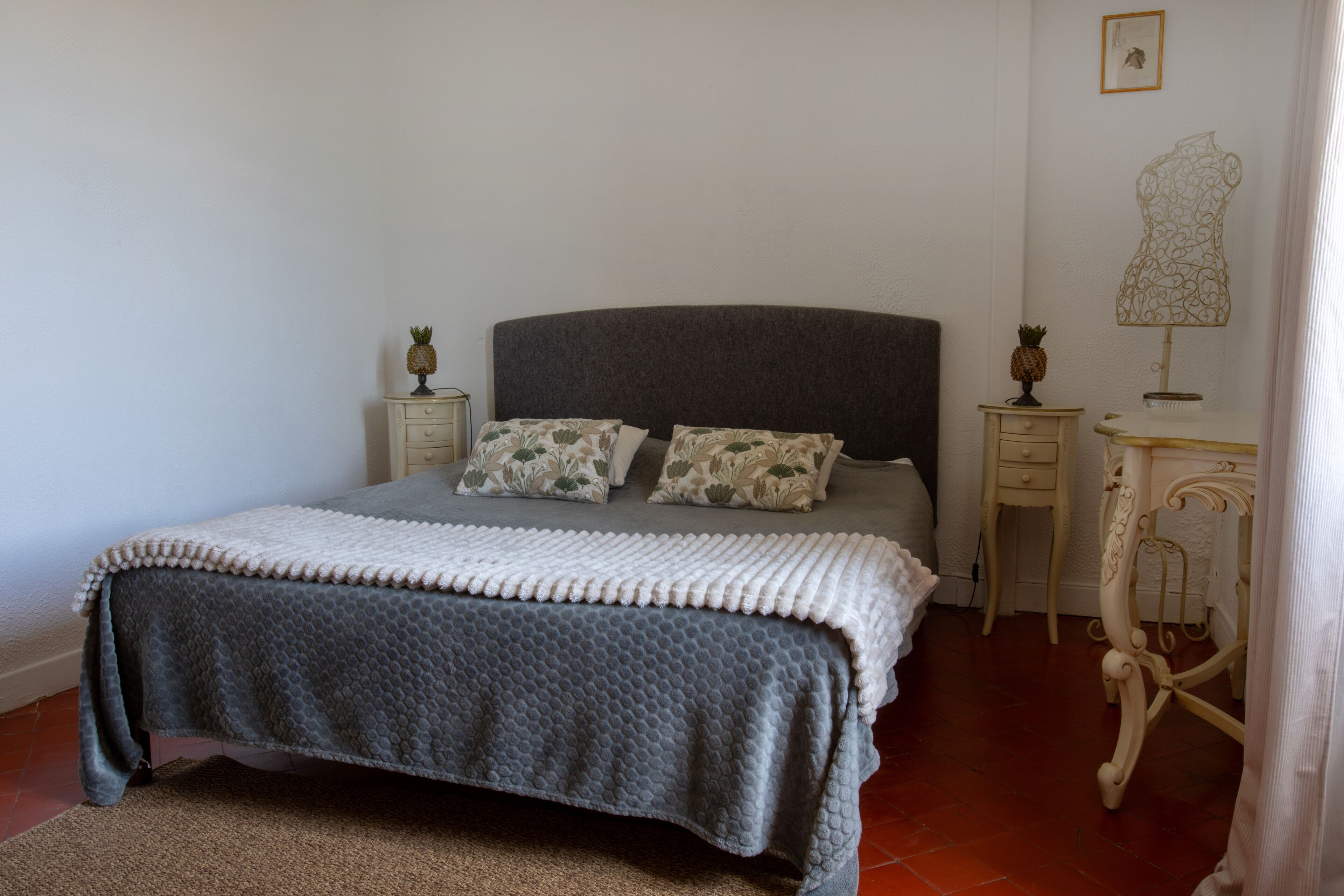 Un lit bien fait, avec une tête de lit grise, deux oreillers assortis, une couverture blanche et grise, et un petit ananas décoratif posé sur la table de chevet.