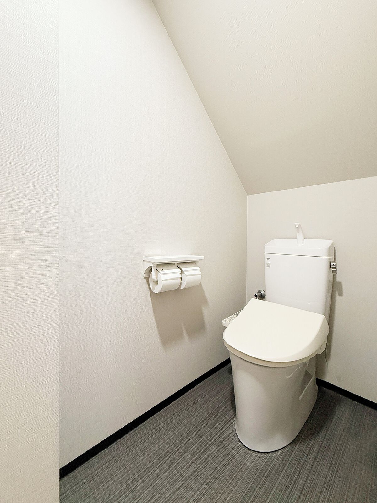A small, modern bathroom with a white toilet, a toilet paper holder, and a grey floor.