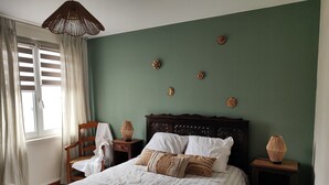 Un dormitorio con cama, mesita de noche, silla, ventana con cortinas y adornos en la pared.