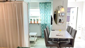 A compact living space with a bed, a dining table with chairs, a small white table, and a mirror. There are potted plants on a shelf and a window with patterned curtains. - 4 Star Holiday Home in Laholm (Skummesloevsstrand)