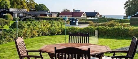 An outdoor wooden deck with a table and chairs, overlooking a well-maintained lawn and a row of houses.