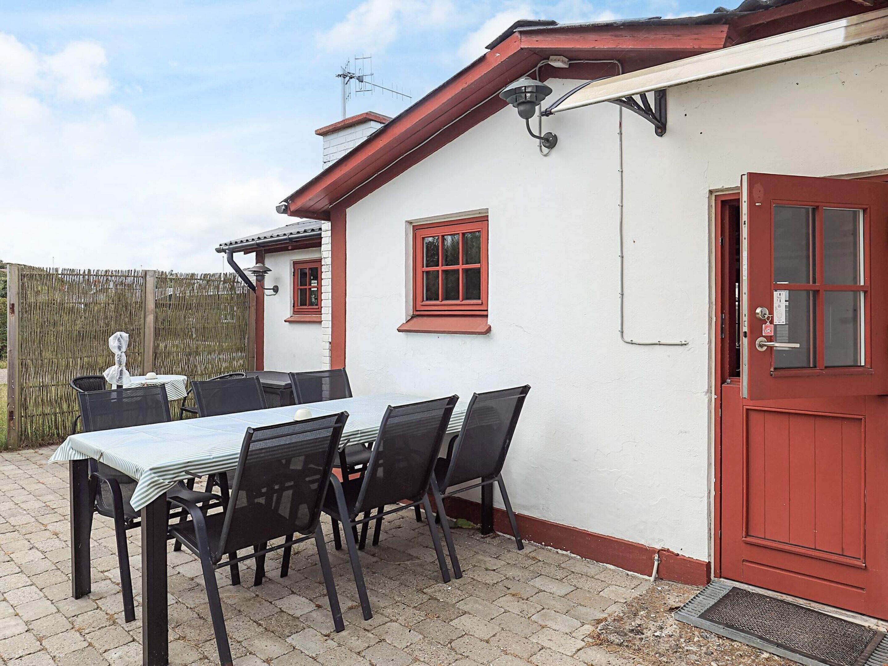 Une terrasse avec une table et des chaises, une porte rouge et un mur blanc avec des fenêtres.