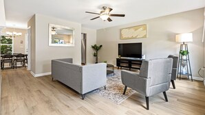 A modern living room with a ceiling fan, grey sofas, a flat-screen TV, and a dining area with a chandelier.