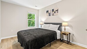 A bedroom with a bed, two bedside lamps, a window with greenery, and artwork on the wall.