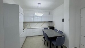 A modern kitchen with white cabinets, a marble countertop, and a dining table with chairs. - Rana E Hedhun Apartment (Lezha)