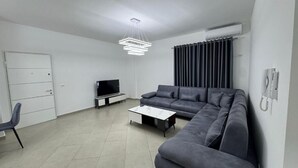 A modern living room with a sectional sofa, a flat-screen TV, a coffee table, and a pendant light. - Rana E Hedhun Apartment (Lezha)
