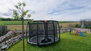 Un trampolín con red de seguridad en un patio trasero con borde de piedra, un tobogán azul y una valla de madera.