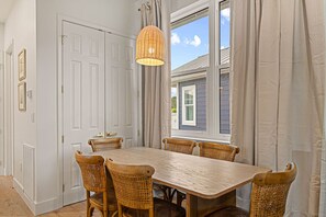 A dining room with a wooden table and wicker chairs, a hanging light fixture, and a view of a house through a window with curtains.