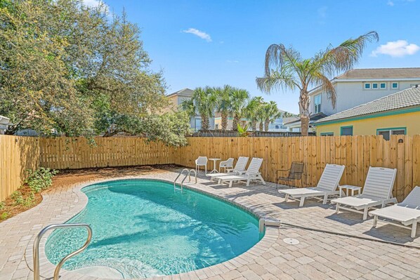 A swimming pool with a wooden fence, lounge chairs, and palm trees.