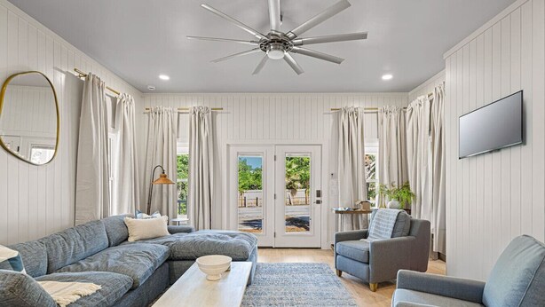 A spacious living room with a ceiling fan, a large flat-screen TV mounted on the wall, and a view through the glass doors to the outside. - 5BDR Renovated Home~Private Pool + Games (Destin)