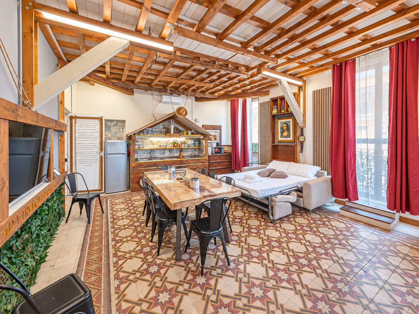 A cozy room with a wooden ceiling, a dining area with chairs, a bed, and a window with red curtains.