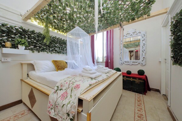 A bedroom with a canopy bed, a bedside table with a red cloth, and a decorative mirror on the wall.