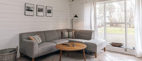 Un salon avec un canapé d’angle gris, une table basse ronde, un lampadaire et une grande fenêtre avec des rideaux.