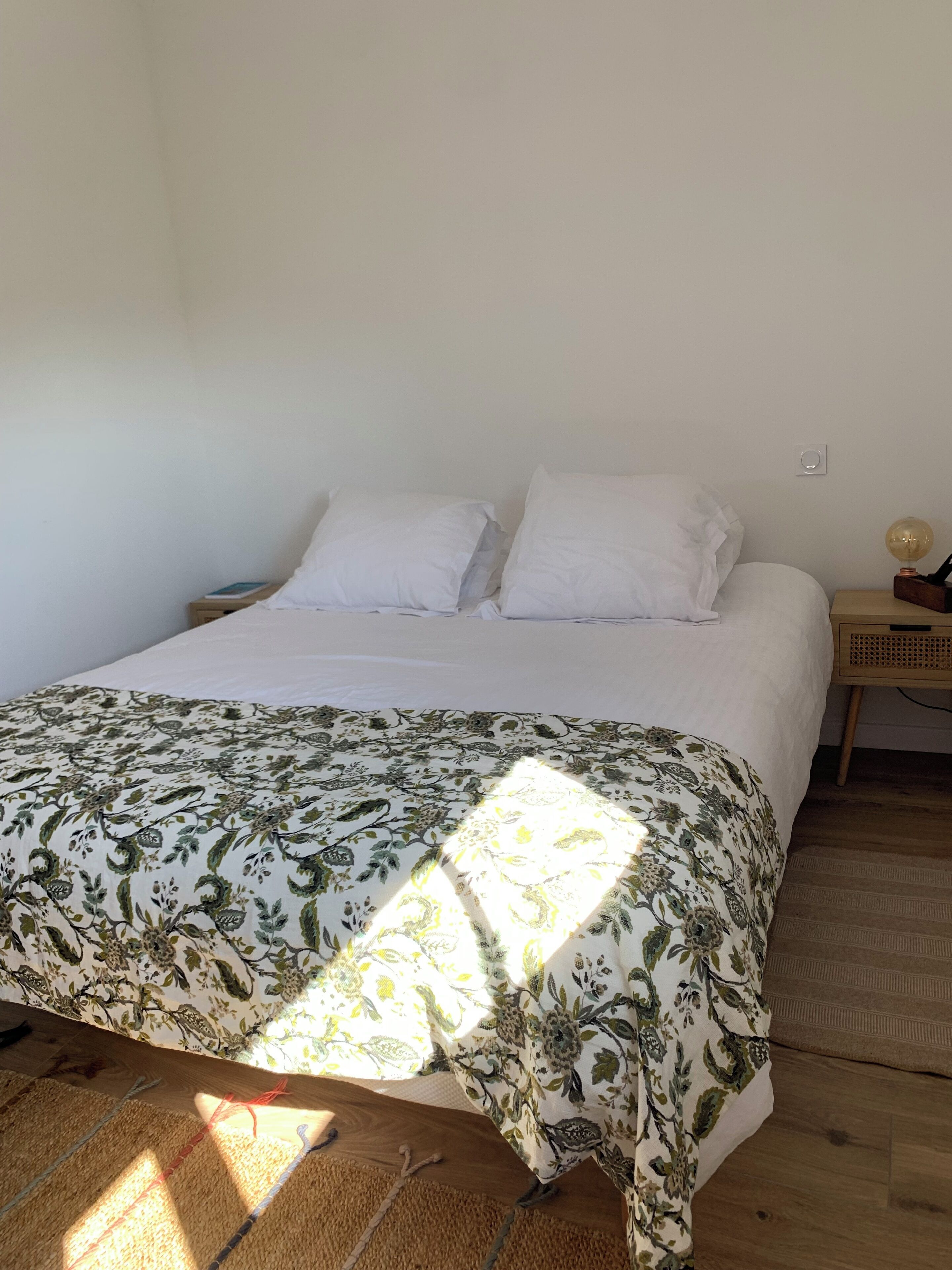 Un lit avec une couvre-lit à motifs floraux, une table de chevet avec une lampe et une prise électrique murale.