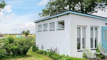 A small, single-story building with a white exterior and a blue door. It features a small patio with two chairs and a table.