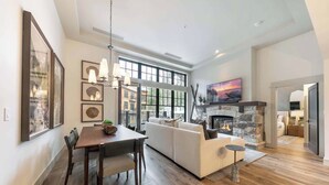 A spacious living room with a fireplace, a dining area with a wooden table and chairs, and a large window providing natural light. - Catamount Lodge 101 - 2 Bedroom (Truckee)