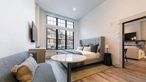 A modern living room with a sofa, a round table, a bed with a headboard, a TV mounted on the wall, and a large window with a view of buildings. - Catamount Lodge 101A -  Studio (Truckee)