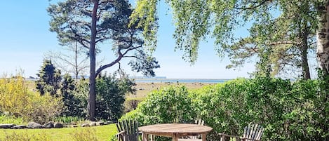 An outdoor wooden deck with a table and chairs, surrounded by greenery and trees, overlooking a beach.