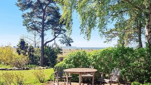 A wooden deck with a round table and four chairs, surrounded by greenery and trees, overlooking a beach. - 4 Person Holiday Home in Klintehamn (Klintehamn)