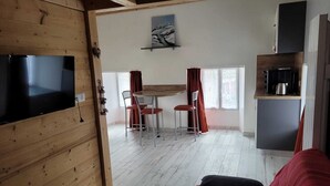 Ein gemütliches Zimmer mit Holzbalkendecke, einer roten Couch, einem an der Wand montierten Fernseher, einer Essecke mit Tisch und Stühlen sowie einer Küchenzeile mit Kühlschrank und Wasserkocher.