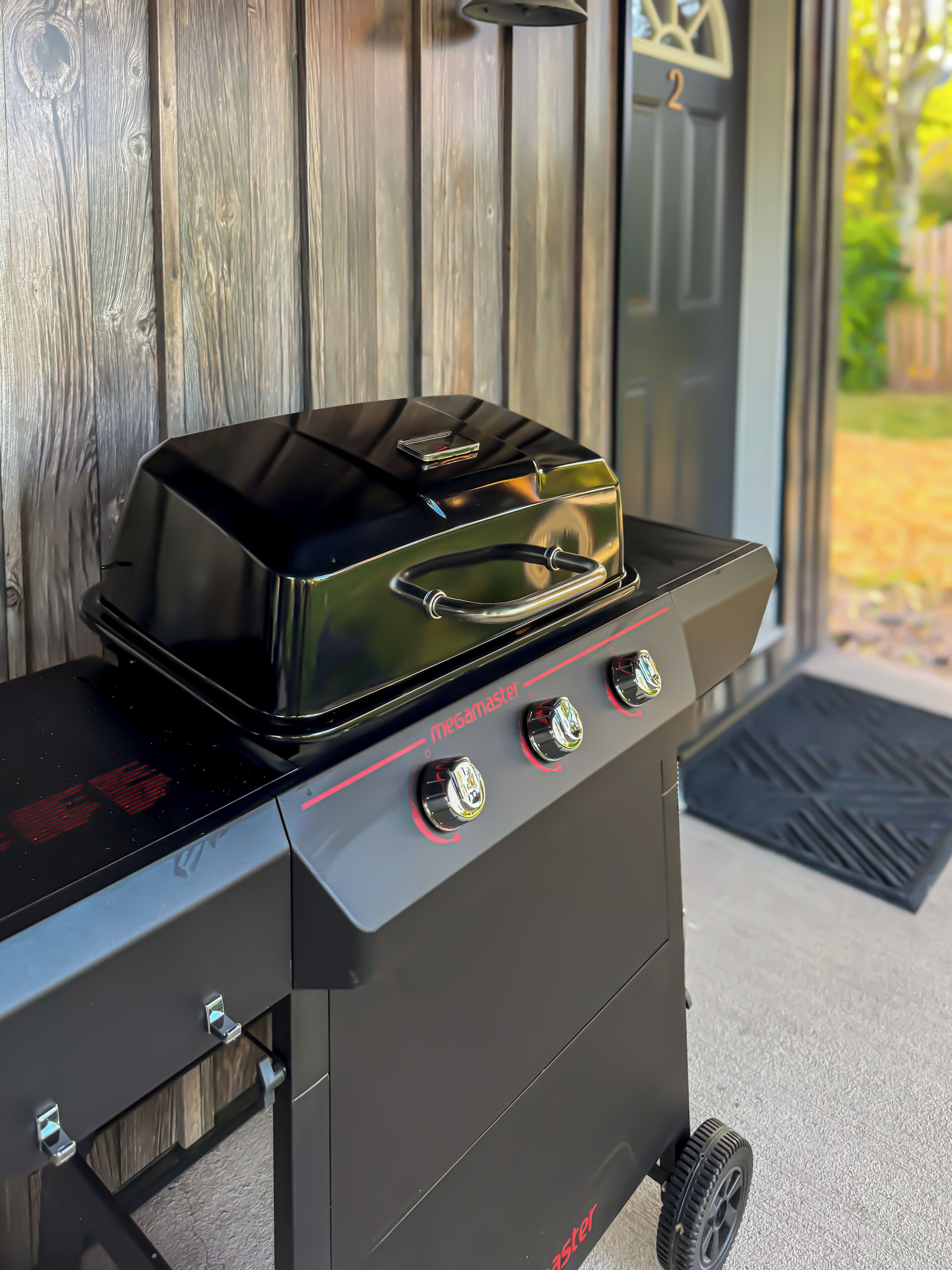 Una parrilla exterior negra con tres quemadores y una pantalla digital, ubicada frente a una puerta de madera.