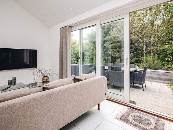 A modern living room with a flat-screen TV, a sofa, and a glass door leading to an outdoor patio with chairs and a table. - 4 Star Holiday Home in Blokhus (Blokhus)