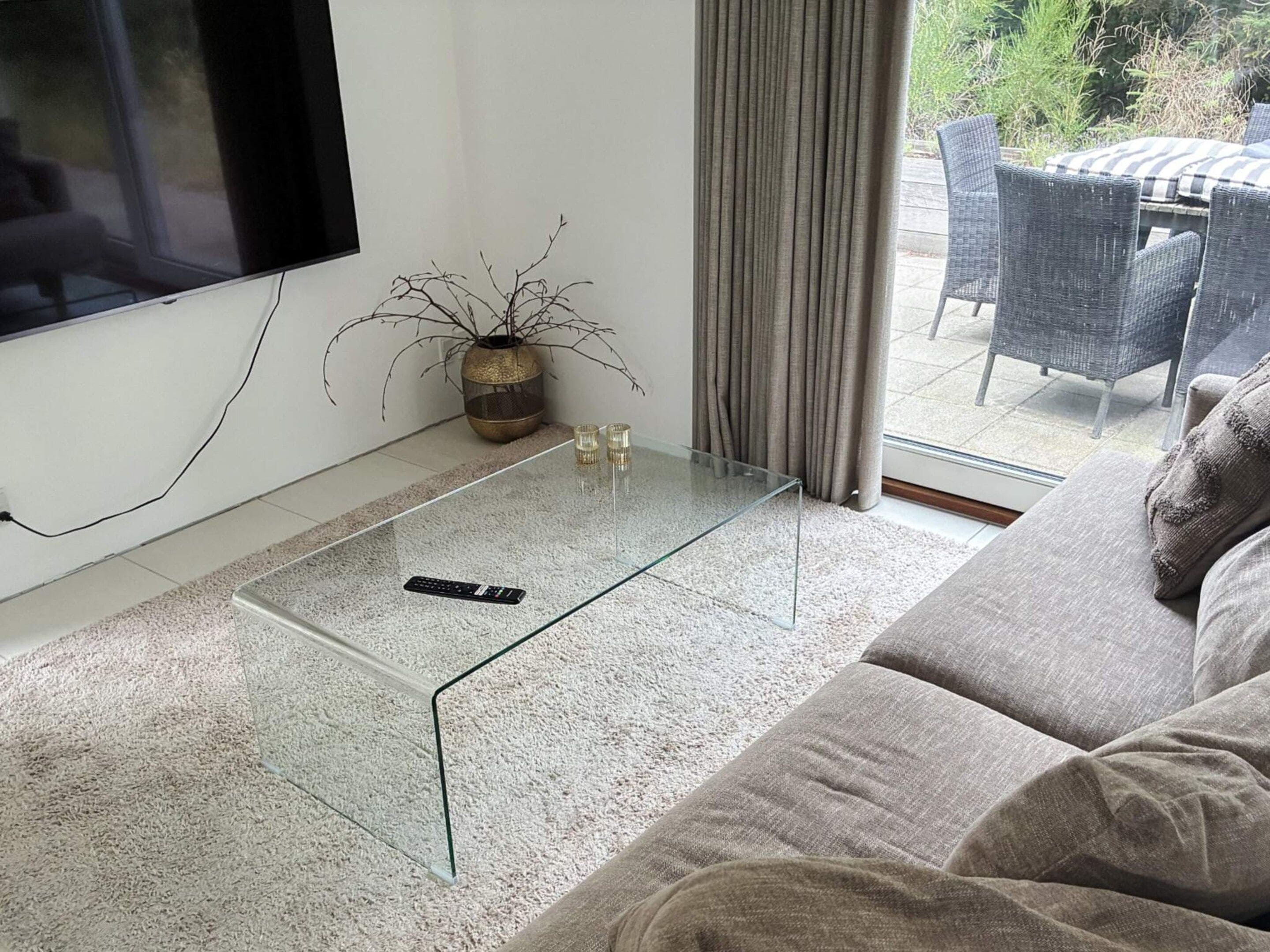 A modern living room with a glass coffee table, a sofa, a TV mounted on the wall, and a view of the outdoors through a glass door.