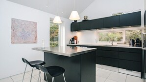 Una cocina moderna con una isla central, taburetes negros tipo barra, iluminación colgante y un amplio ventanal.