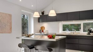 Una cocina moderna con una isla central, dos taburetes de bar, iluminación colgante y un amplio ventanal.