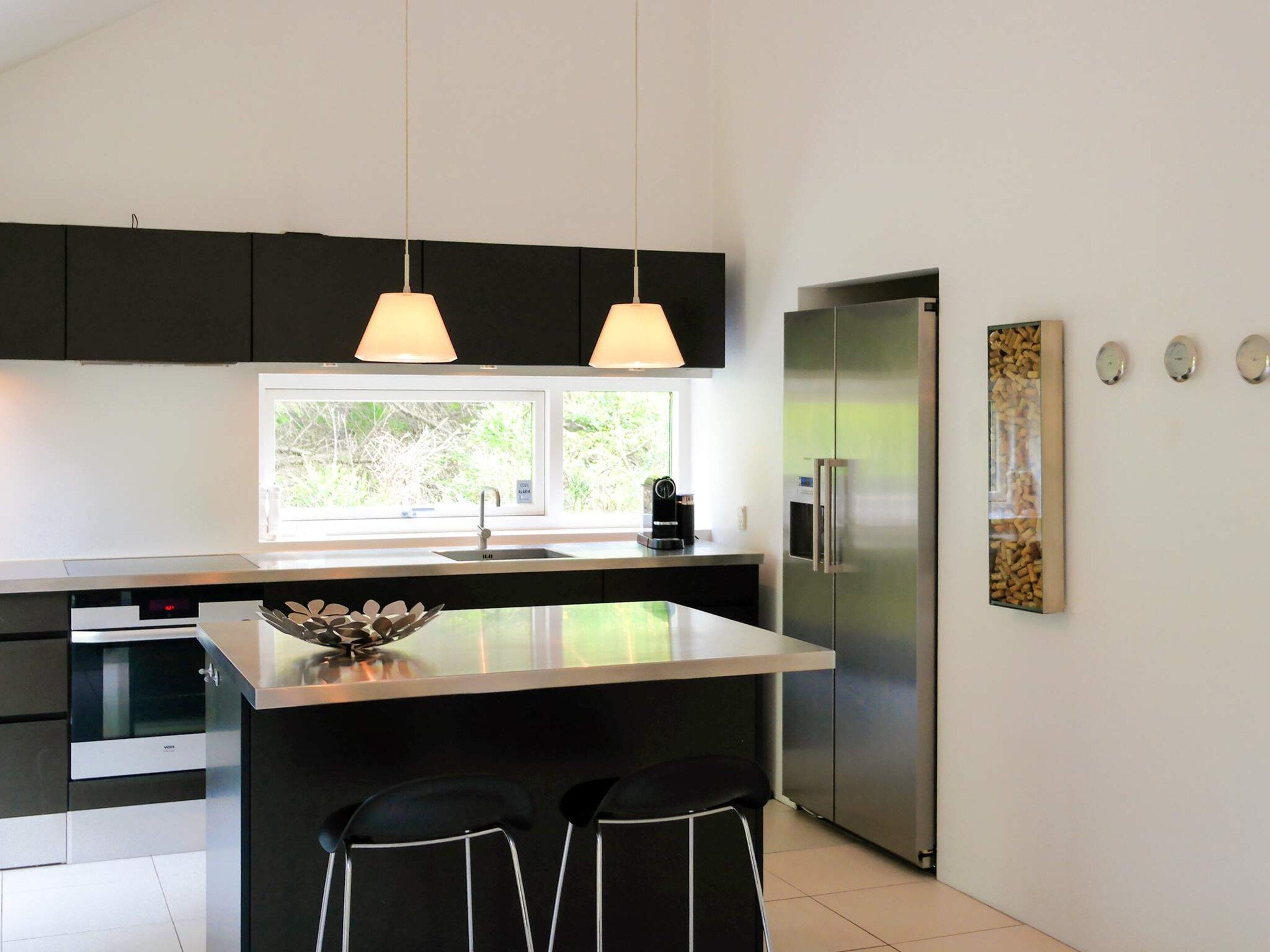 A modern kitchen with a central island, stainless steel appliances, pendant lighting, and a window with a view of greenery.
