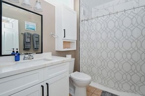 Un bagno con lavandino bianco, specchio e servizi igienici. Si nota una tenda da doccia con motivo geometrico e un porta asciugamani sopra il lavandino.