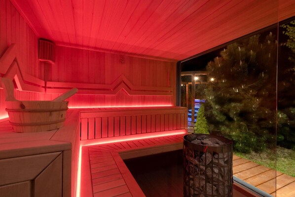 A sauna with wooden benches and a stone container, likely for ashes, with a view of a Christmas tree outside.