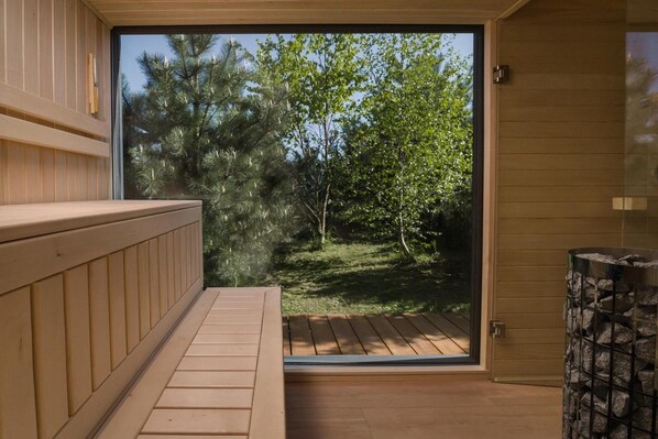 A sauna room with a wooden bench and a glass door leading to a garden with trees and a wooden deck.