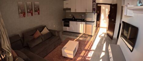 A modern living room with a grey sofa, a white coffee table, and a fluffy rug. There is a kitchen area with white cabinets and a built-in oven. A flat-screen TV is mounted on the wall.
