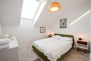 Un dormitorio con tragaluces, cama con cabecera verde, lámparas de noche, mesita de noche con lámpara y un cuadro en la pared.