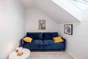 A room with a blue sofa, two yellow pillows, a small white table with a candle, and artwork on the walls.